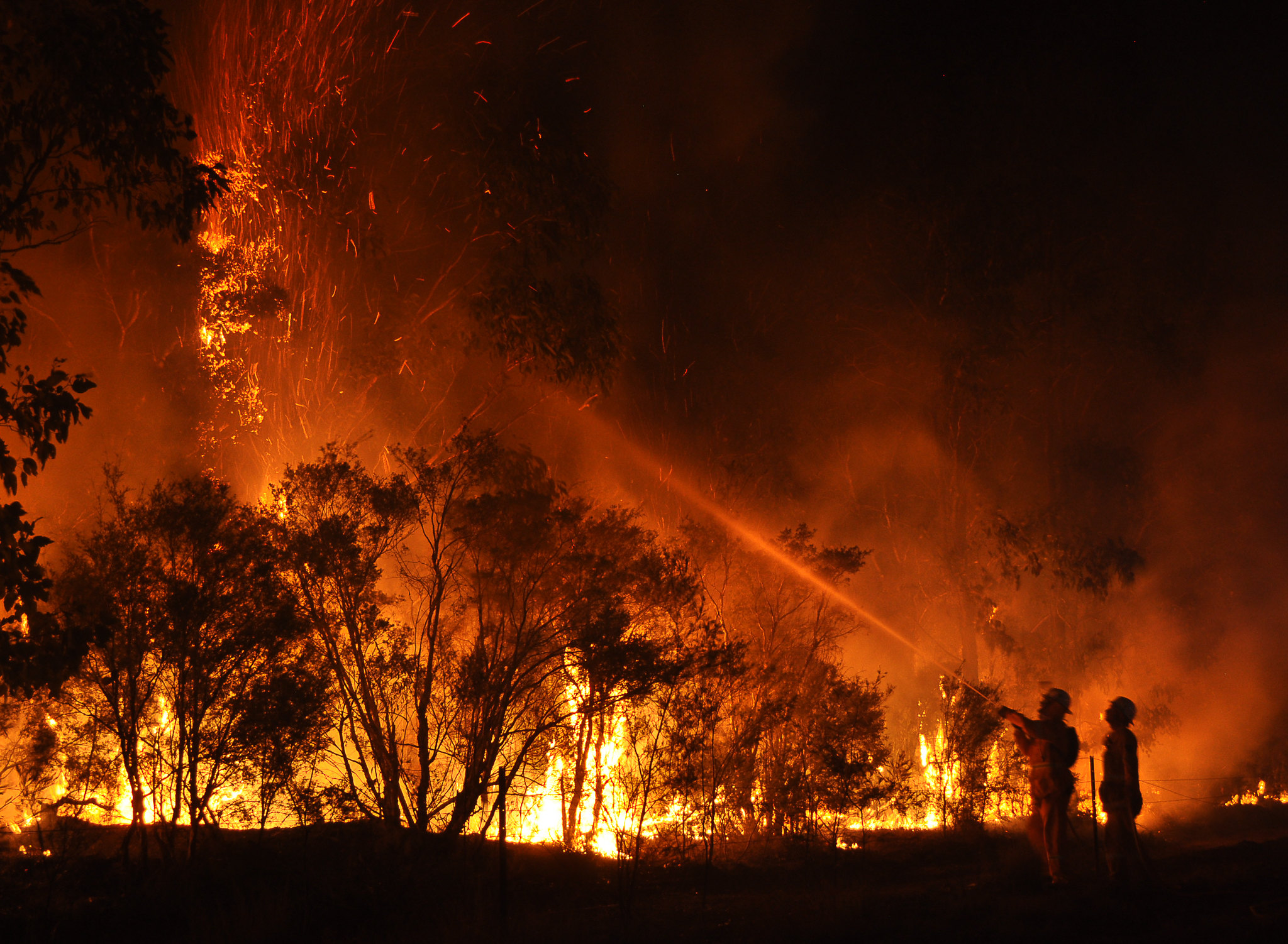 Blazing wildfire burns against a night sky as firefighters spray water on the flames